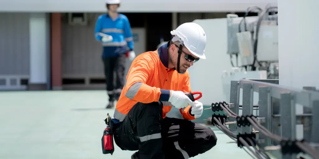 Techniker in Sicherheitskleidung arbeitet an Elektrokabeln, trägt Helm und Handschuhe.