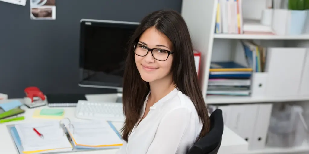 Eine junge Frau mit Brille und langen Haaren lächelt am Schreibtisch im Büro.