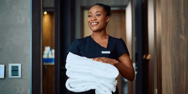 Eine lächelnde Frau in Arbeitskleidung hält frische weiße Handtücher in einem modernen Hotelflur.