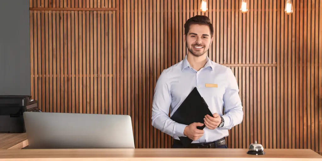 Hotelrezeptionist lächelt, hält Klemmbrett, Holzvertäfelungshintergrund, Empfangsglocke, moderne Beleuchtung.