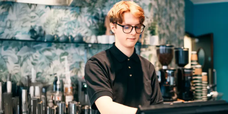 Junger Mann an der Kasse eines Cafés mit tropischem Hintergrund und Kaffeemaschinen.